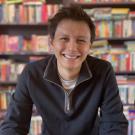 Photo of Ben smiling behind colorful bookshelf. 